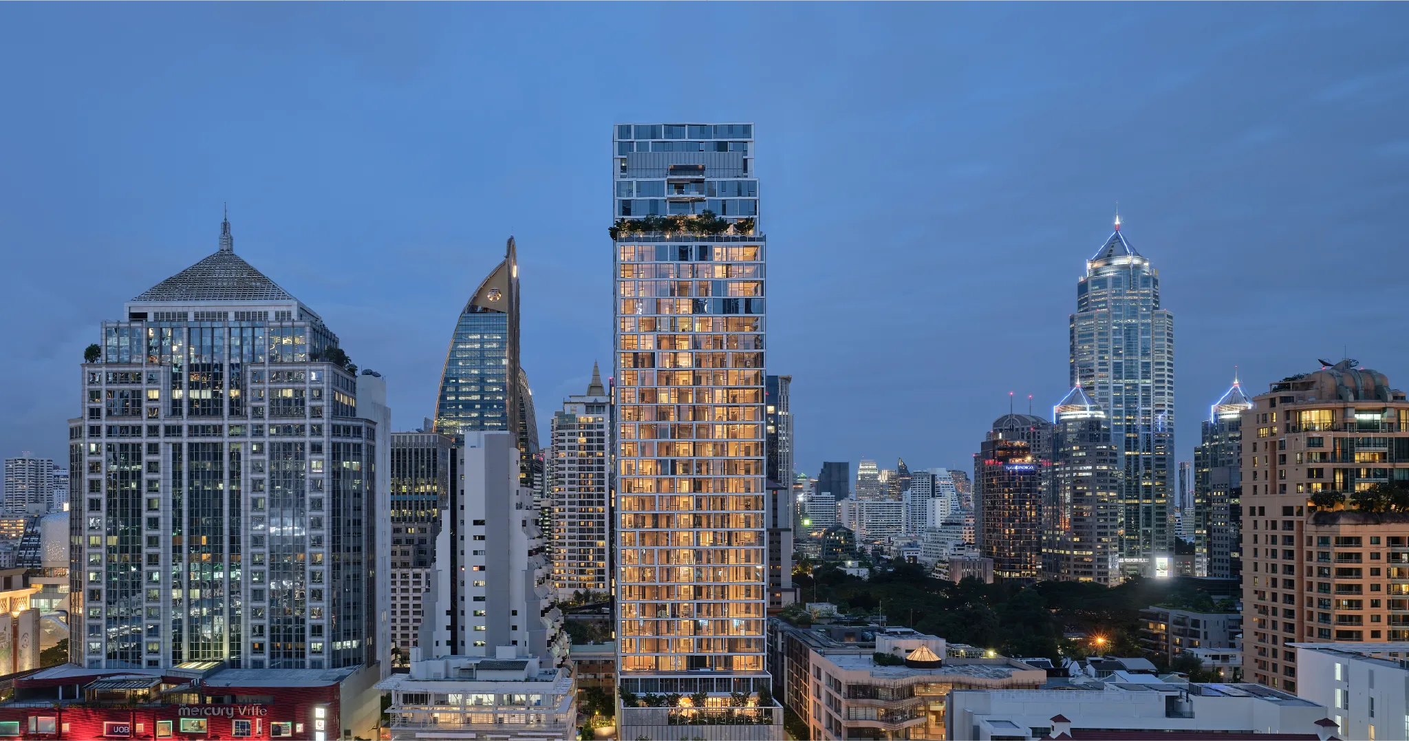 อาคารสูงของคอนโดใกล้รถไฟฟ้าอย่างสโคปตอนกลางคืนที่มีตึกต่าง ๆ ในกรุงเทพอยู่รอบข้าง 